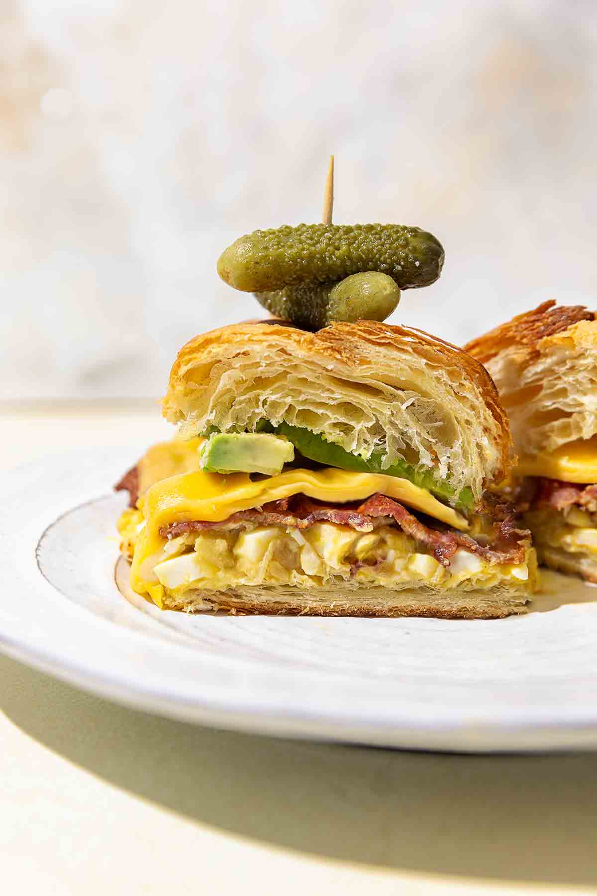A halved croissant sandwich with egg salad, bacon, cheese, avocado, and small pickles on a white plate.