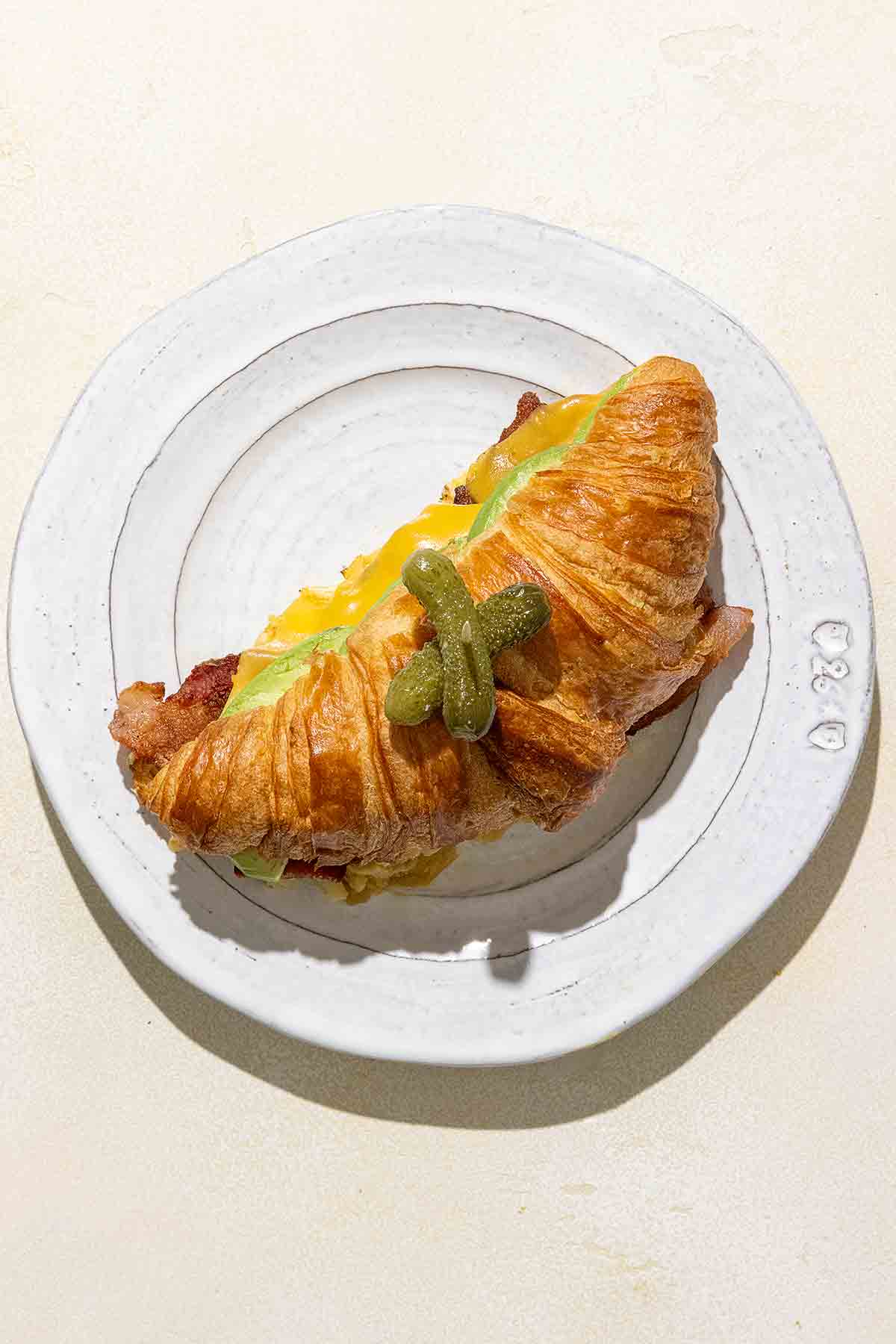 A croissant sandwich topped with two baby pickles on a white plate.