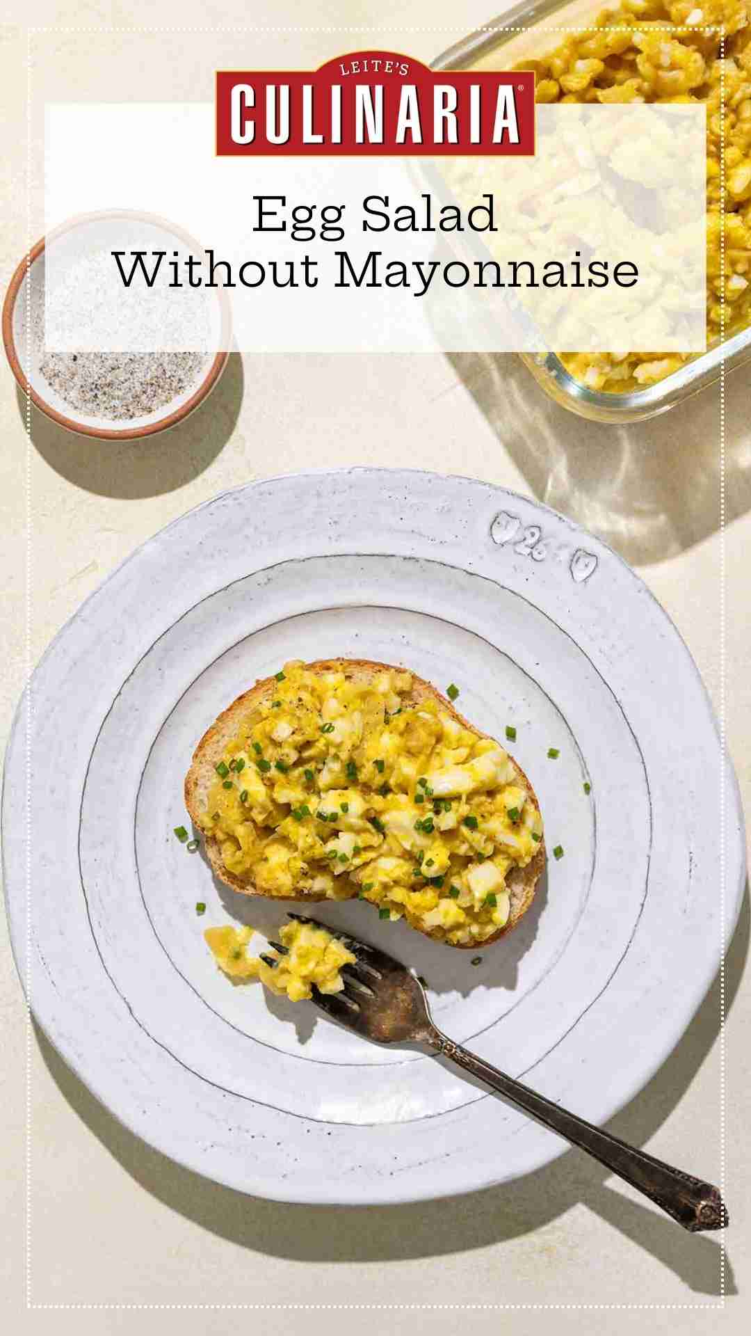 An open-faced egg salad sandwich topped with minced chives on a white plate with a bowl of salad and a dish of salt and pepper nearby.