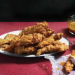 Homemade chicken strips piled on a white plate with dishes of barbecue sauce and ranch nearby.