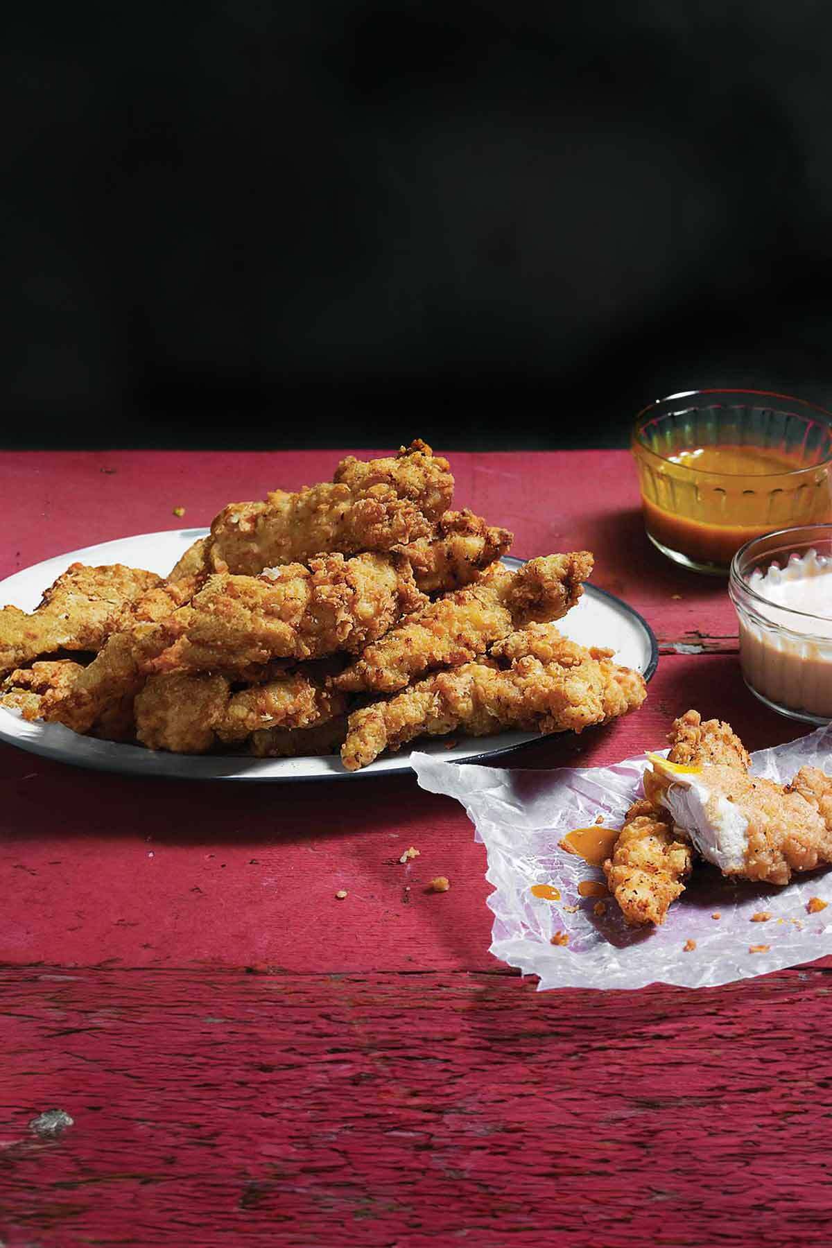 Homemade chicken strips piled on a white plate with dishes of barbecue sauce and ranch nearby.