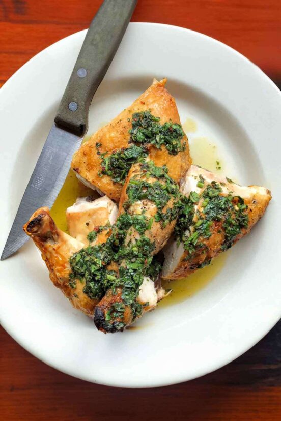 Pieces of roast chicken topped with salsa verde on a white plate, with a knife resting on the side.
