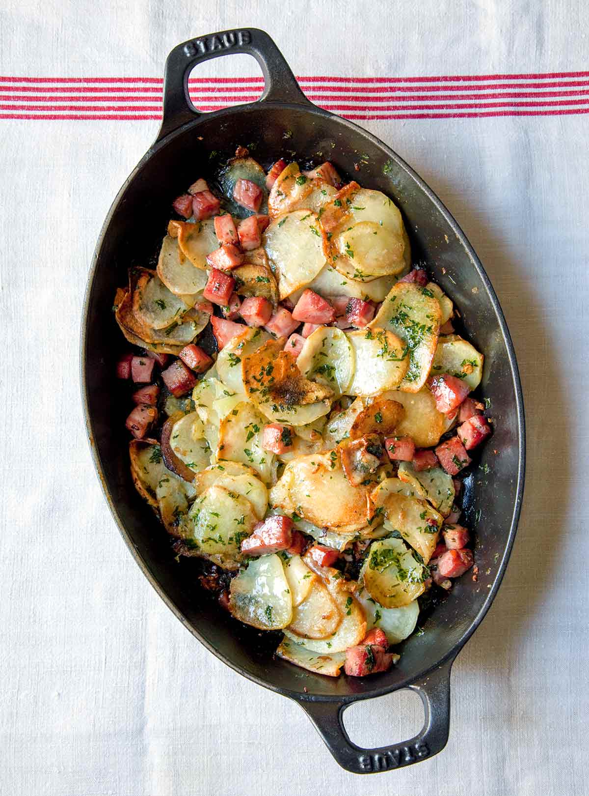 An oval baking dish filled with sliced potatoes, cubed ham, and chopped herbs.