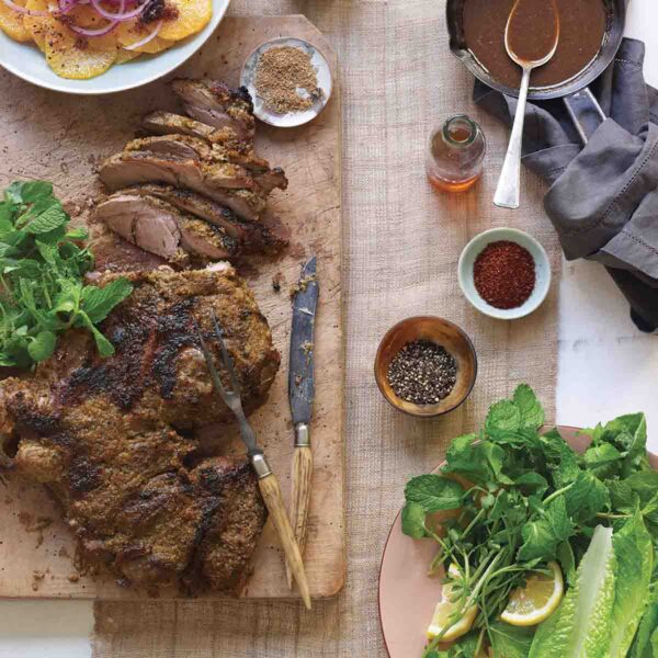 A partially carved roast leg of lamb on a wooden cutting board with spices, fresh herbs, and citrus salad surrounding it.
