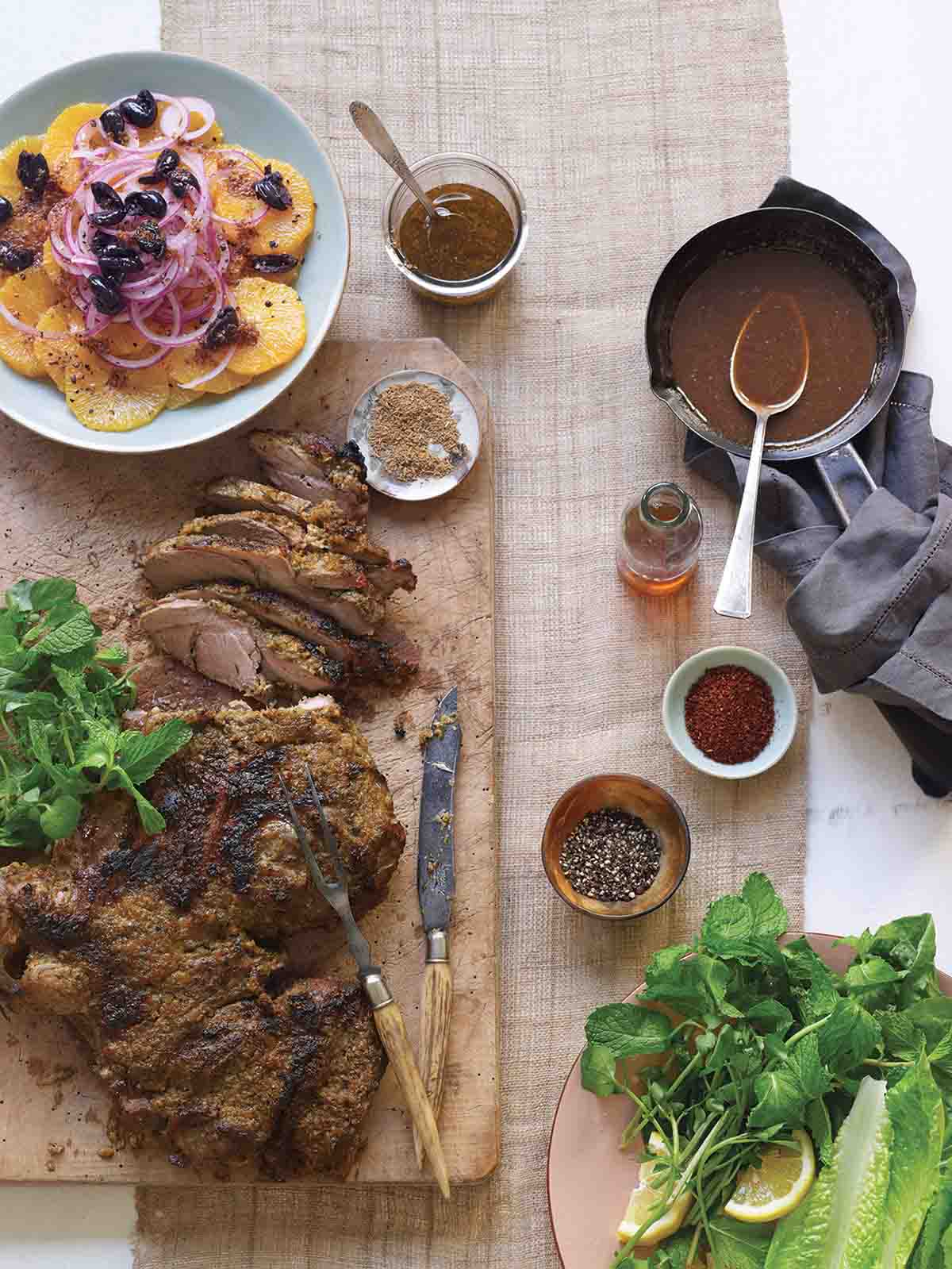 A partially carved roast leg of lamb on a wooden cutting board with spices, fresh herbs, and citrus salad surrounding it.