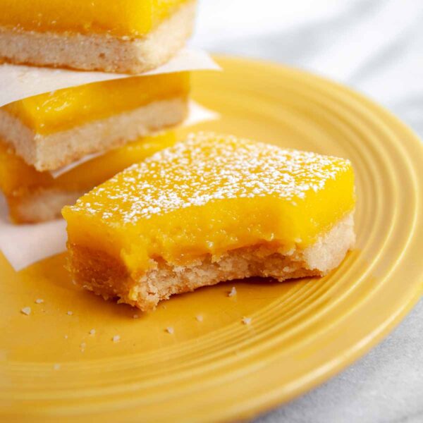 Yellow plate with four lemon bars sprinkled with powdered sugar, one with a bite take out of it.