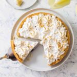 A lemon meringue tart in a white dish, with one slice on a plate beside it and one more being lifted out.