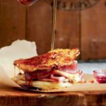 Maple syrup being poured over a monte cristo sandwich that is on a piece of parchment paper.