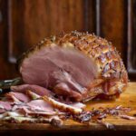 A partially carved mustard-glazed ham, cross-hatched and studded with cloves on a wooden cutting board.