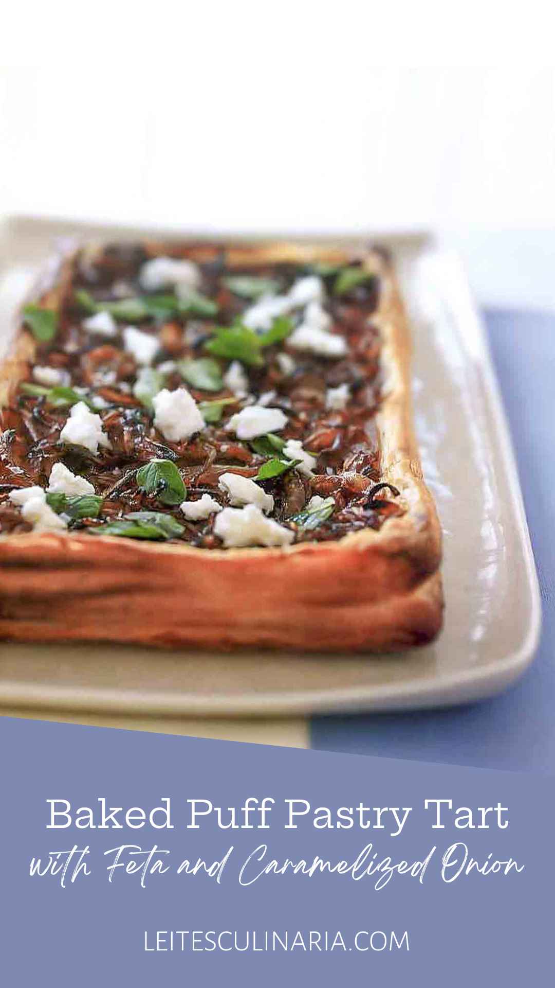 A whole puff pastry tart topped with caramelized onions, feta cheese, and oregano leaves on a rectangular platter.