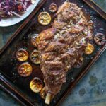 A whole roast leg of lam on a rimmed baking sheet with charred lemon slices scattered around it.