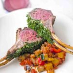 Two chops from a roasted rack of lamb with parsley, Dijon, and chives on a white plate with a cooked zucchini and tomato mixture.