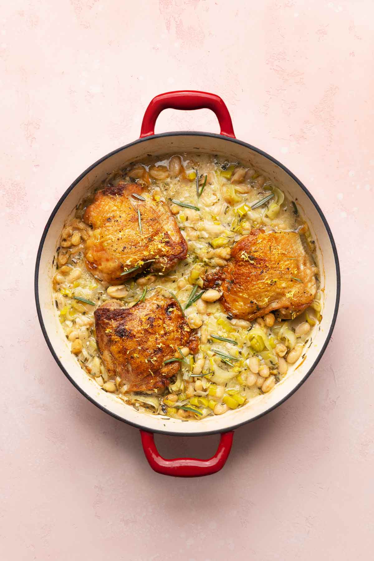 A deep skillet with three chicken thighs on top of a mixture of leeks, rosemary, and cannellini beans.