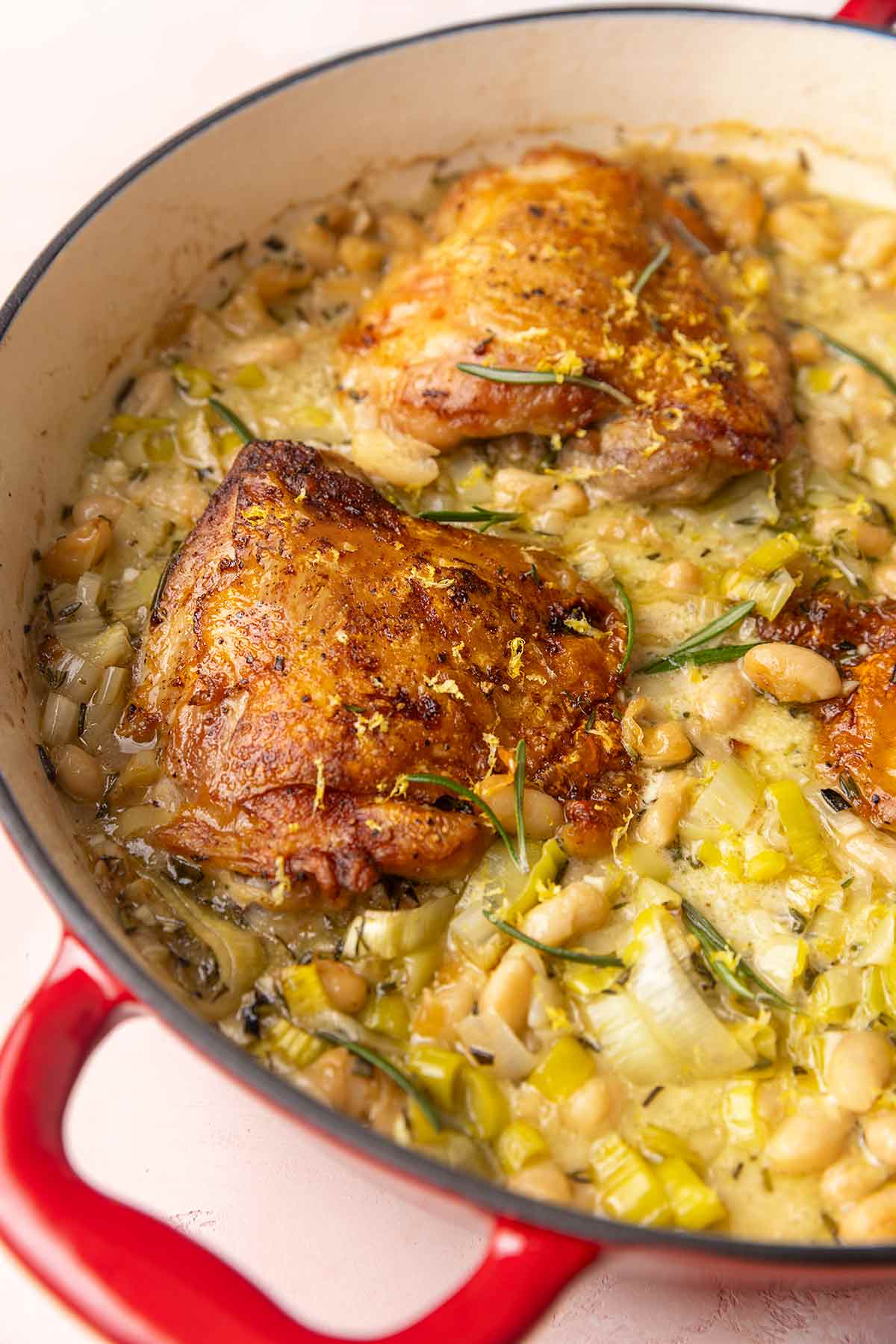 A skillet with three chicken thighs on top of a mixture of leeks, rosemary, and cannellini beans.