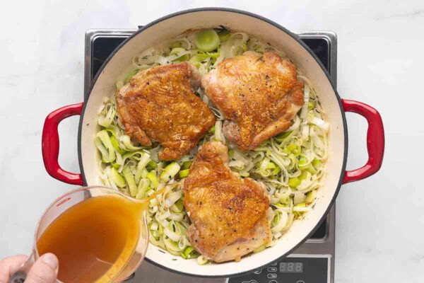A person pouring apple cider over chicken thighs and sliced leeks in a skillet.