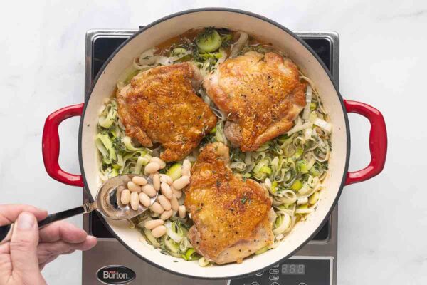 A person spooning cannellini beans into a skillet of chicken thighs and leeks.