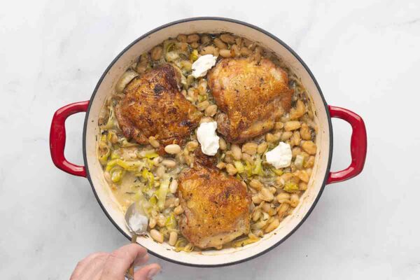 A person stirring sour cream into a skillet of chicken thighs, leeks, and cannellini beans.