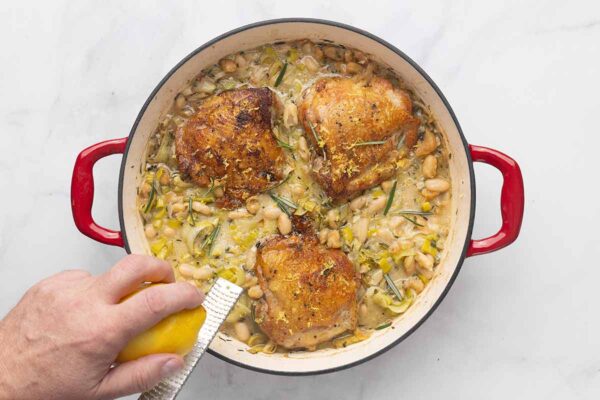 A person zesting a lemon over a skillet of chicken thighs, leeks, cannellini beans, and sour cream.