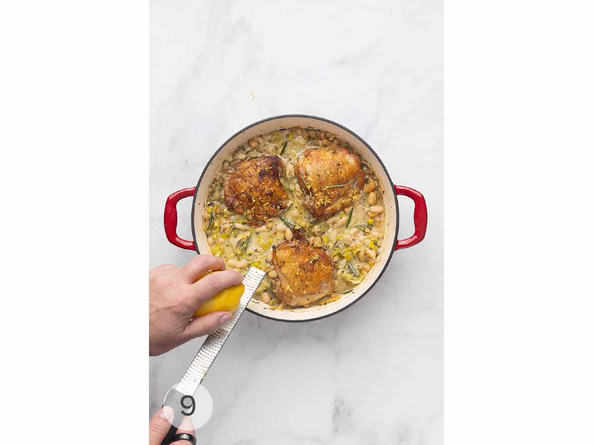 A person zesting lemon into a skillet of chicken thighs, cannellini beans, and leeks.