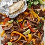 An enamel baking tray filled with sliced peppers, steak strips, onions, lime wedges, cilantro, and a foil packet of tortillas on the side.