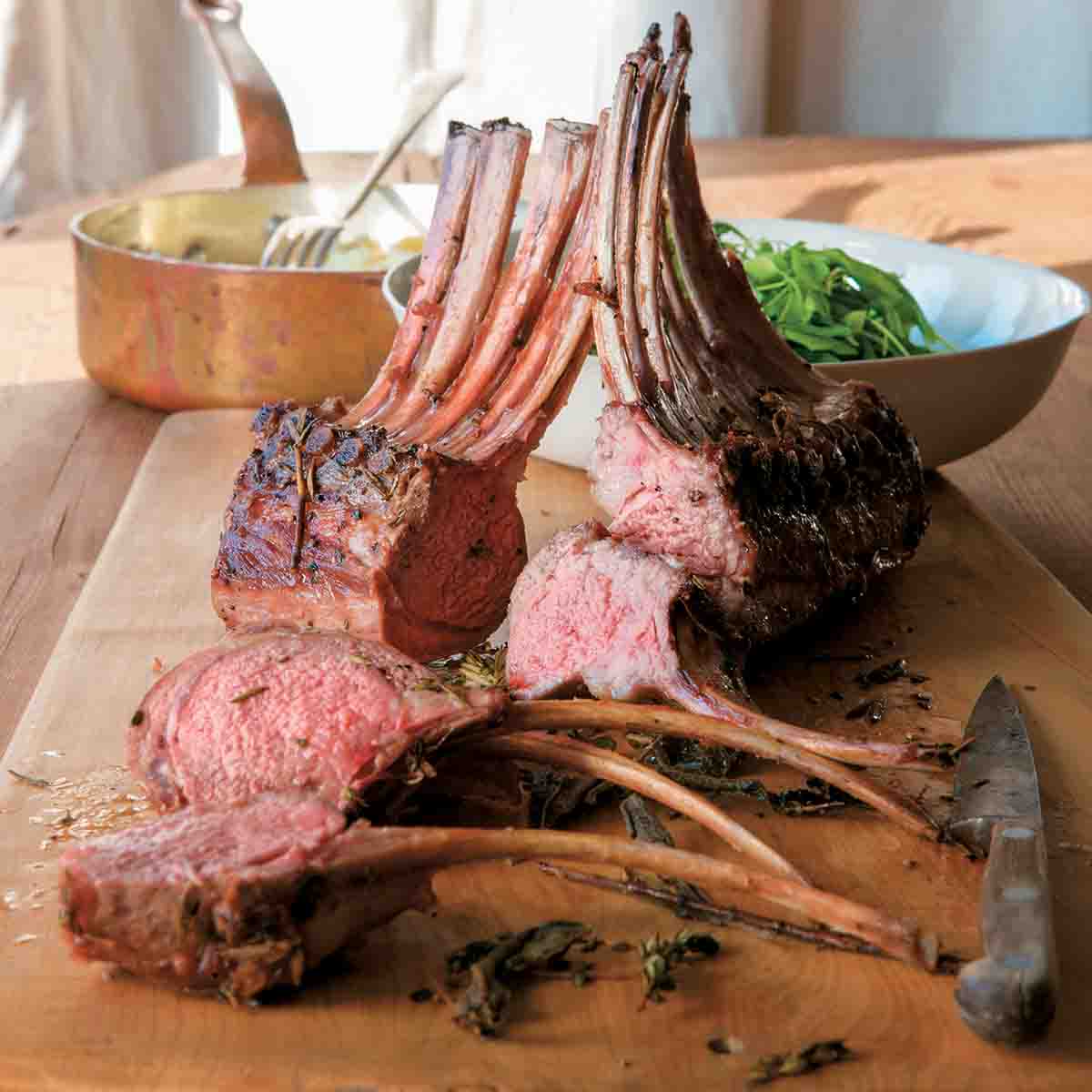 Cutting board with a sliced rack of spring lamb with rosemary, knife, bowl of salad.