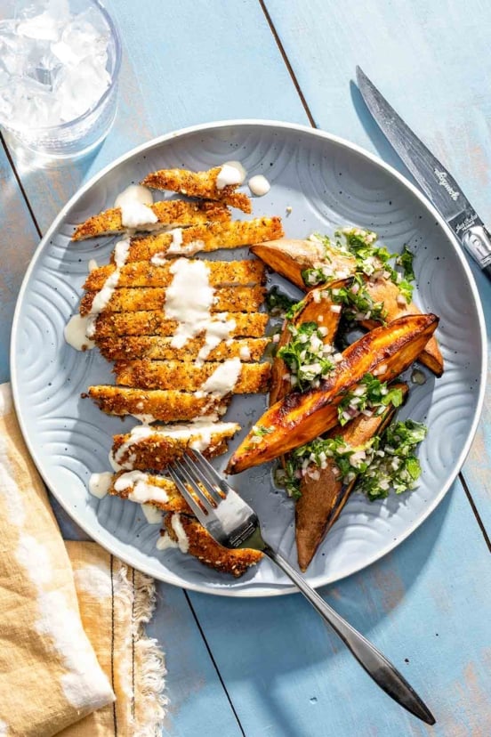 A sliced crispy chicken cutlet drizzled with white sauce and a serving of roasted potato wedges on the side.