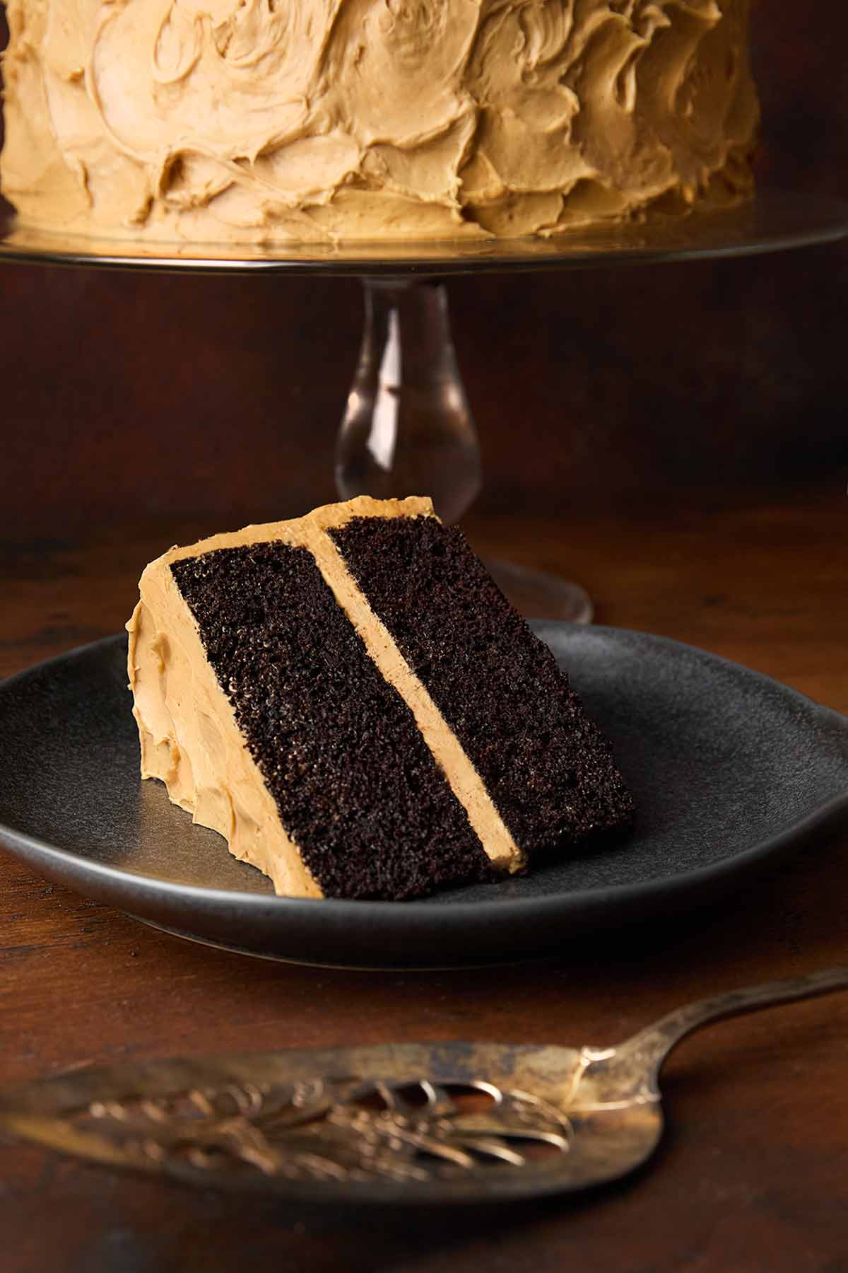 A slice of chocolate caramel cake on a plate, with the rest of the cake in the background.