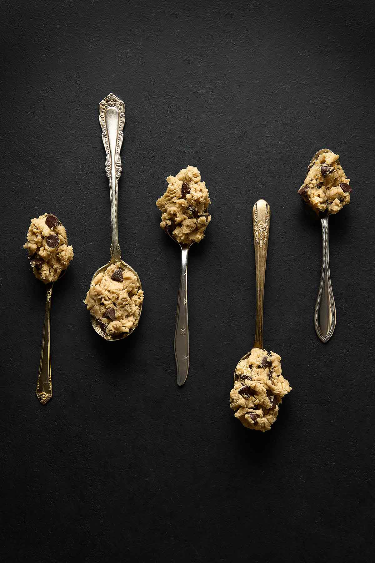 Five silver spoons, each with a scoop of eggless chocolate chip cookie dough.
