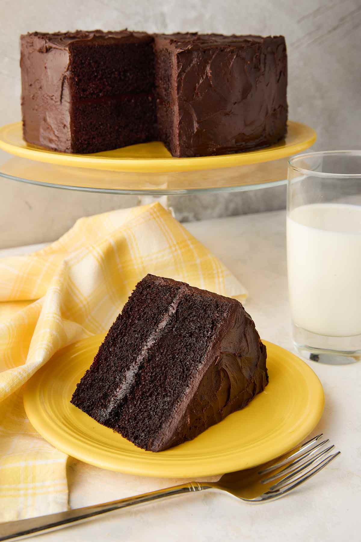 A slice of Hershey's chocolate cake with chocolate frosting on a yellow plate, with the remaining cake on a stand in the background.