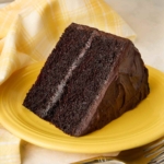 A slice of Hershey's chocolate cake with chocolate frosting on a yellow plate, with the remaining cake on a stand in the background.