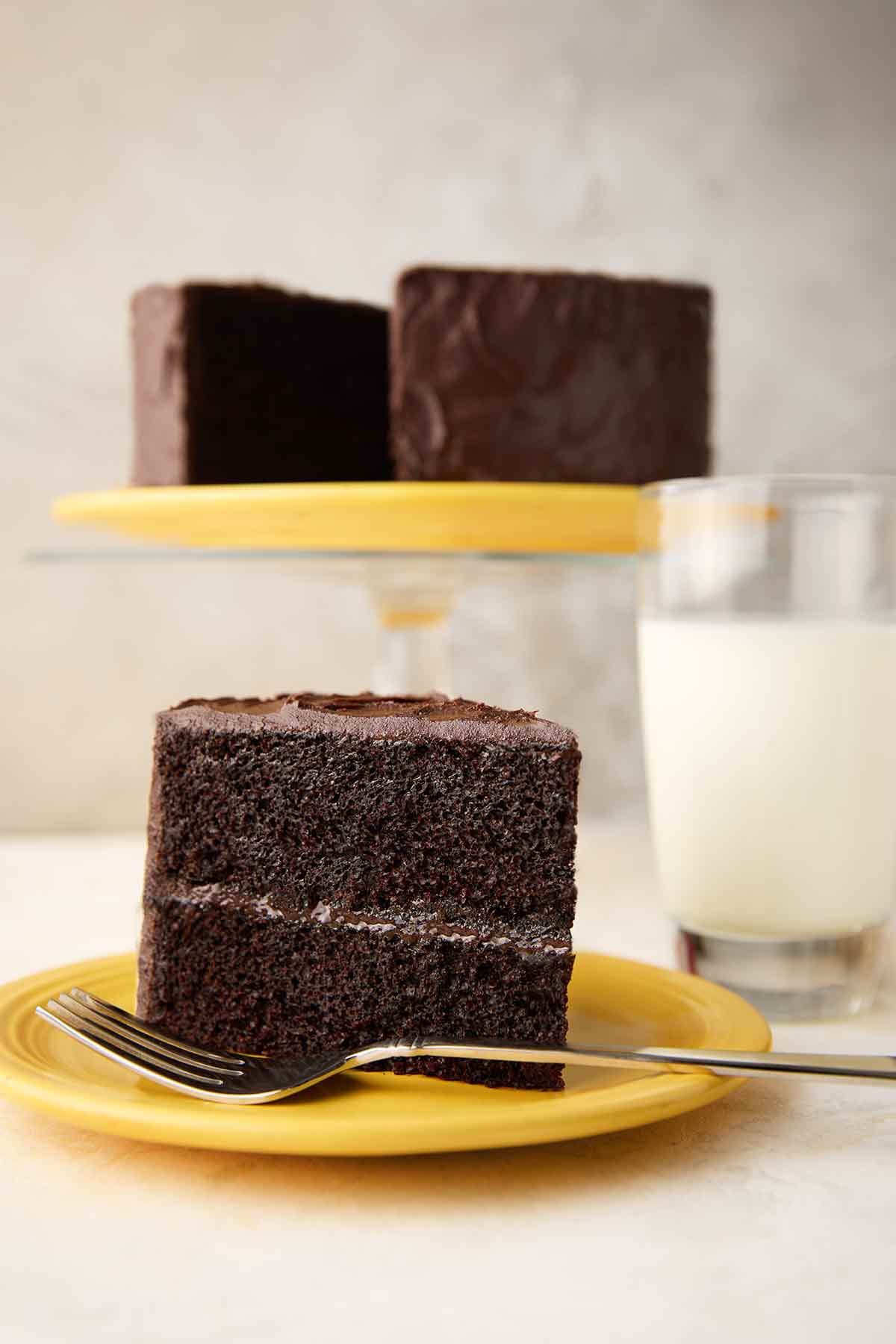 A slice of Hershey's chocolate cake with chocolate frosting on a yellow plate, with the remaining cake on a stand in the background.