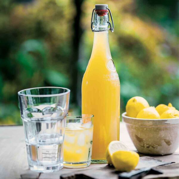 A glass bottle filled with homemade lemonade syrup, a bowl of lemons, and two glasses on a wooden table.