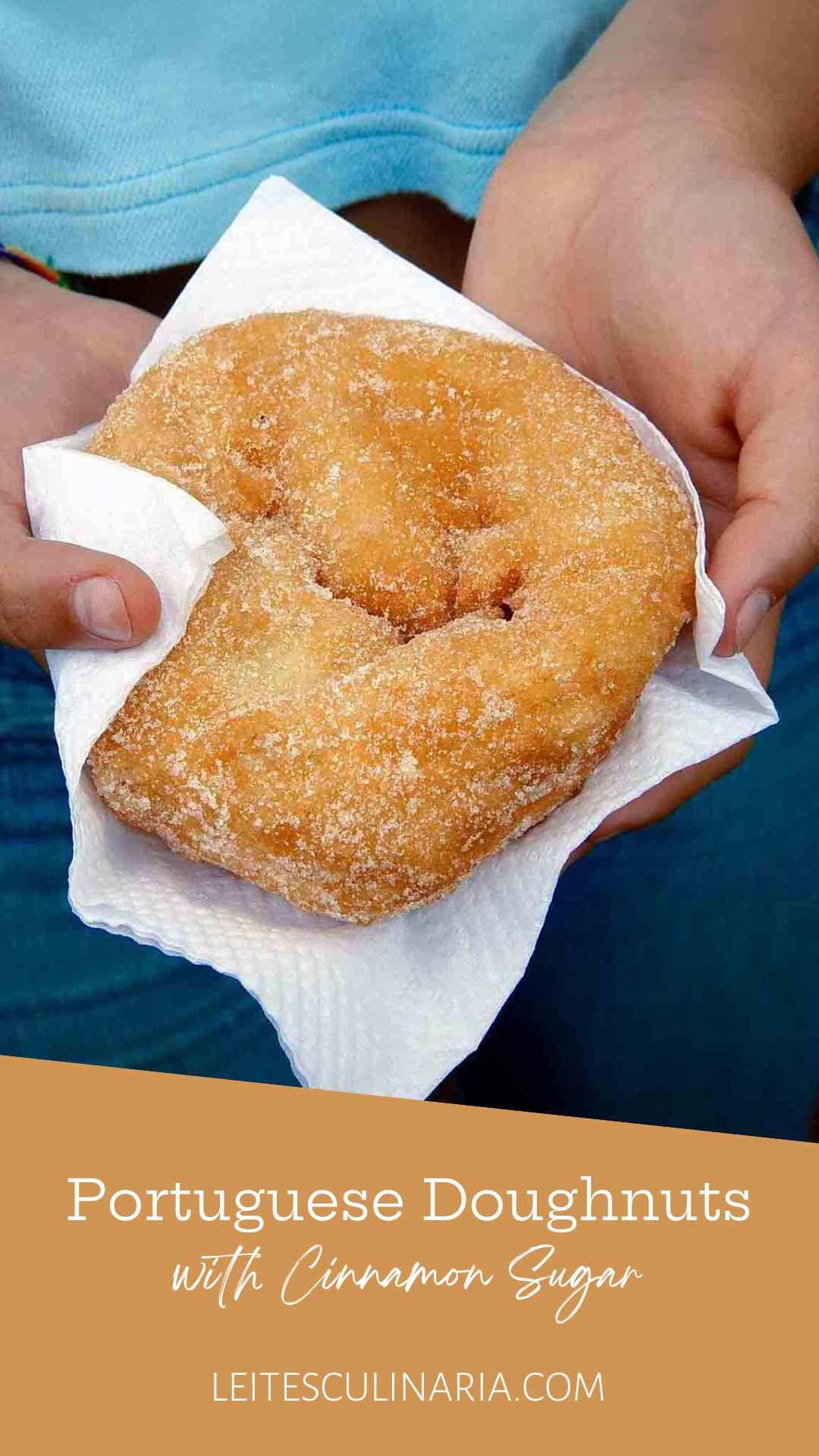 A person holding a fried Portuguese doughnut covered in cinnamon sugar in a paper napkin.
