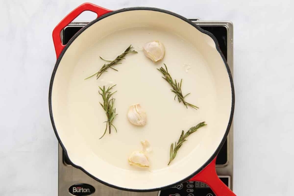 Three garlic cloves and four rosemary sprigs in a skillet on a hot plate.