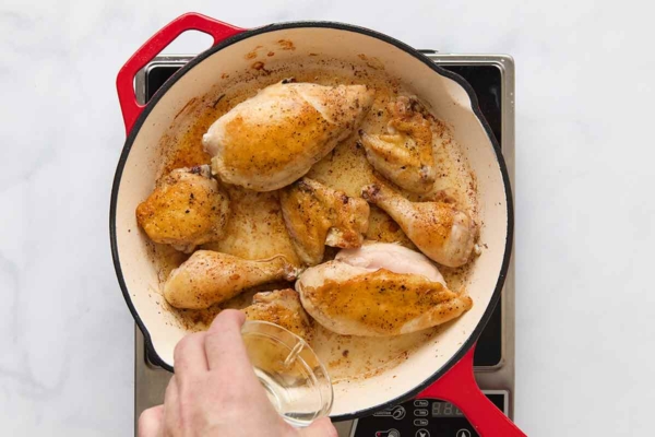 Pieces of chicken being browned in a skillet and a person adding some liquid to the skillet.