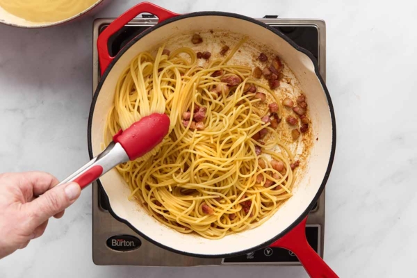 A person tossing spaghetti and crispy cubed pancetta in a skillet.