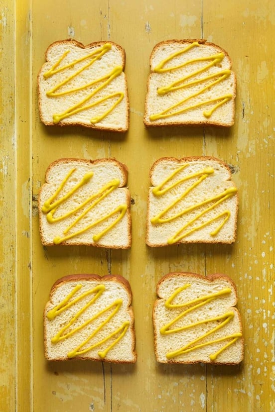 Six slices of white bread with mustard squiggles on each slice.