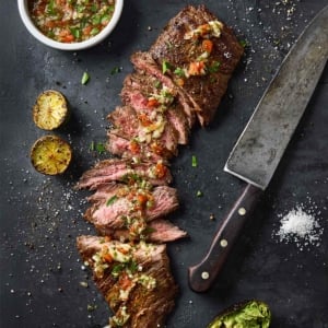 Sliced skirt steak topped with salsa with a bowl of salsa and charred lime halves on the side.