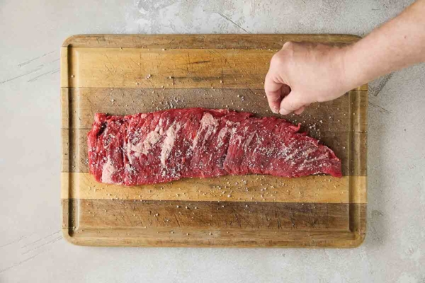 A person seasoning a skirt steak on a cutting board with salt.