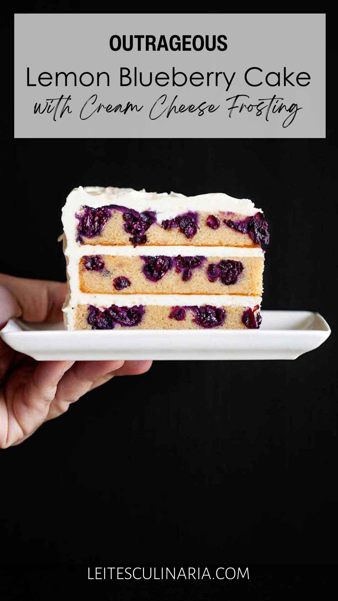 A person holding a slice of three layer blueberry lemon cake with cream cheese frosting on a white rectangular plate.