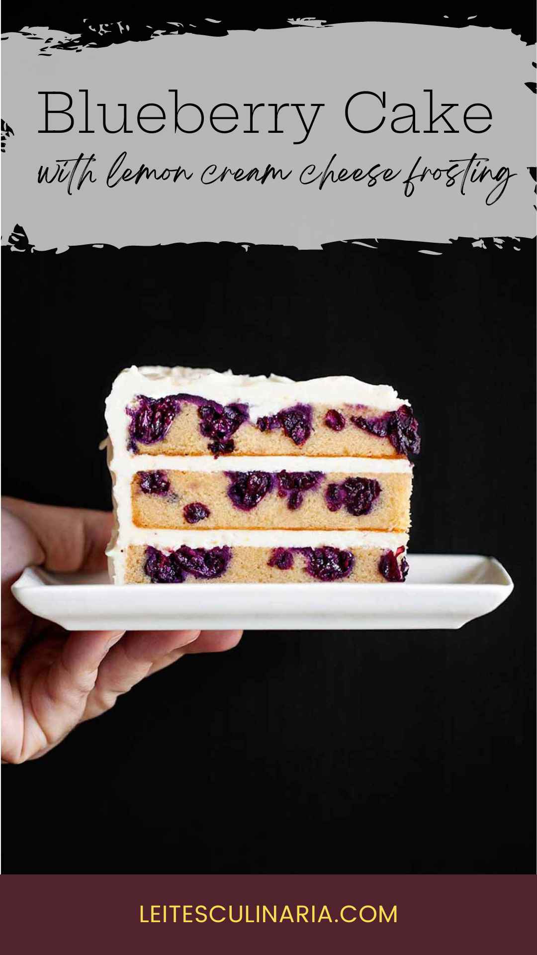 A person holding a slice of three layer blueberry lemon cake with cream cheese frosting on a white rectangular plate.