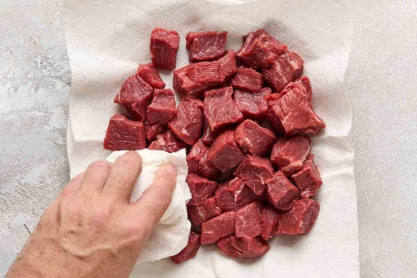 A person patting cubes of beef dry with paper towel.