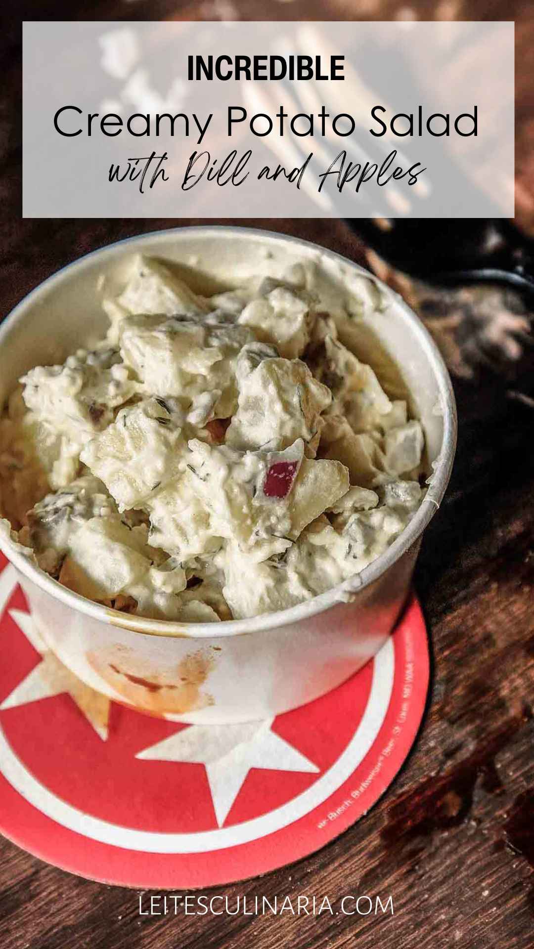 A container of creamy potato salad with apples, red onion, and dill on a red and white coaster.