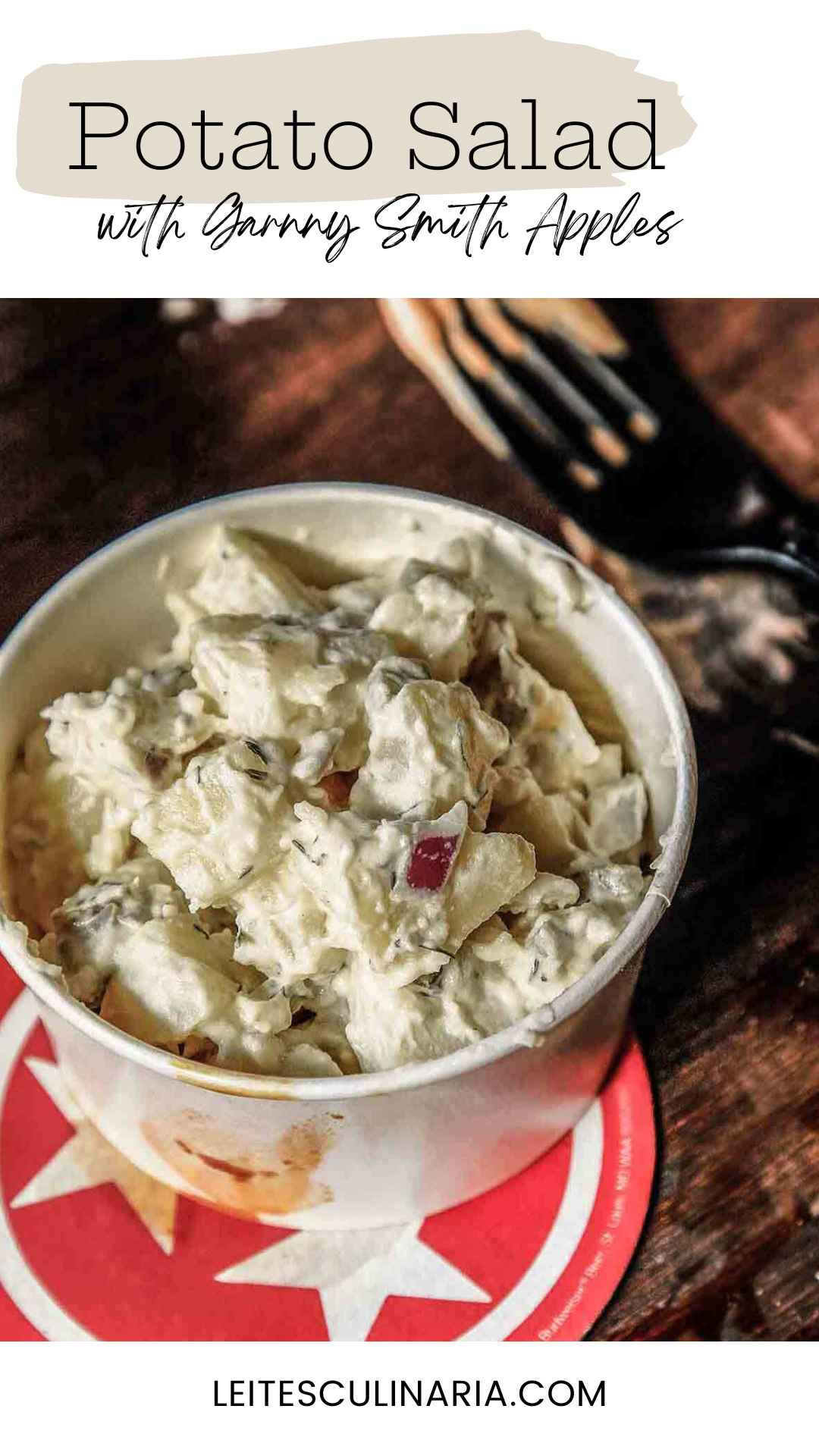 A container of creamy potato salad with apples, red onion, and dill on a red and white coaster.