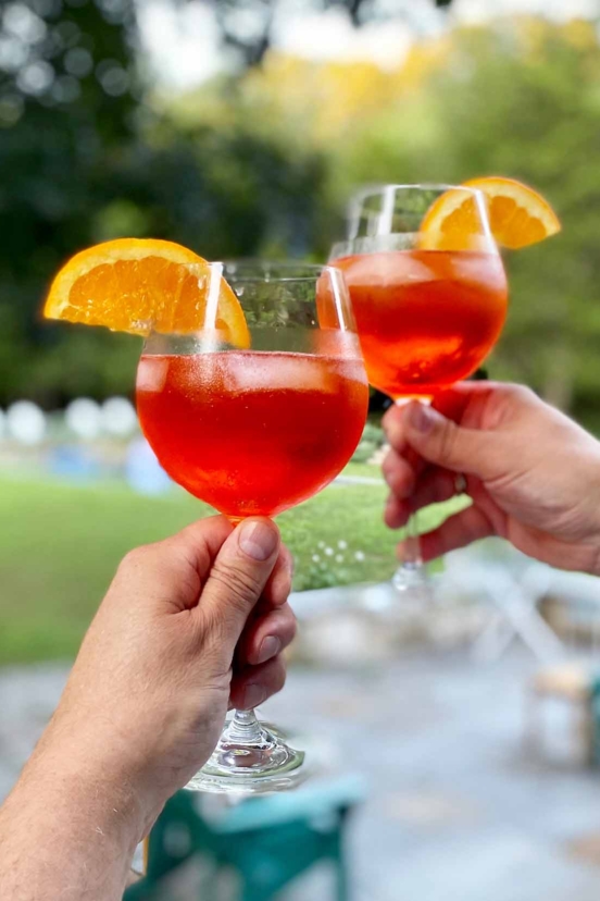 Two people toasting each other with glasses of Aperol spritz, garnished with orange slices.
