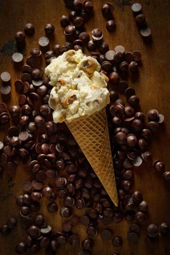 A sugar cone of chocolate chip cookie dough ice cream lying on a bed of chocolate chips.