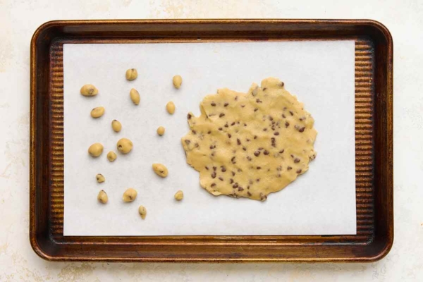 A piece of chocolate chip cookie dough and several small balls of dough on a baking sheet.