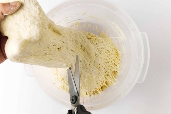 A person cutting a chunk of dough from a large container of bread dough.