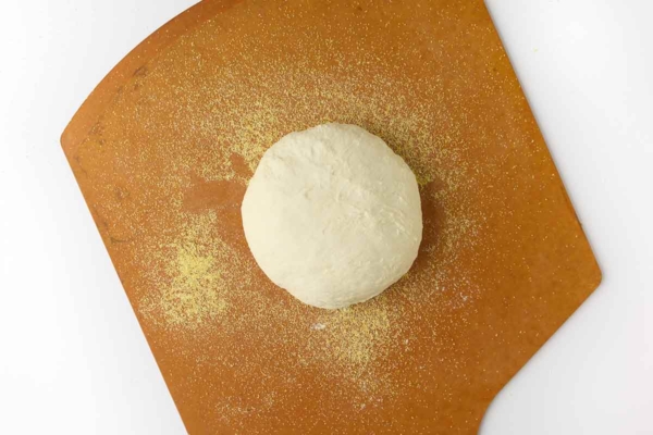A round of bread dough on a pizza peel dusted with cornmeal.