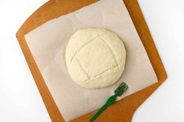 A scored round of bread dough on a sheet of parchment on a pizza peel.